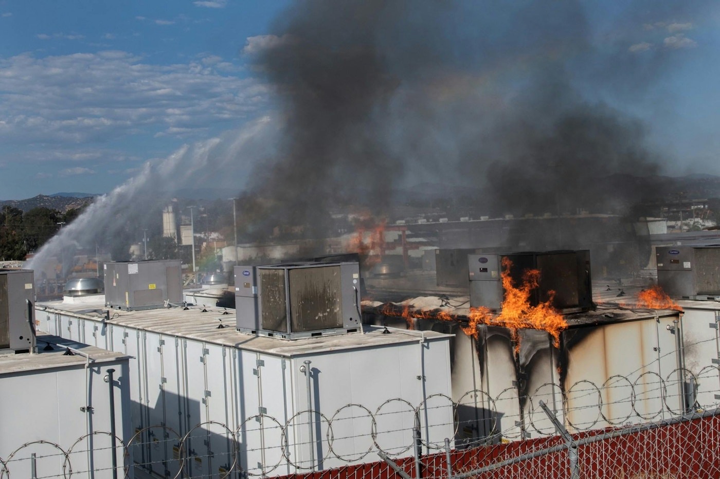 Active flames and thick black smoke rising from a burning lithium-ion battery storage container at the SDG&E Escondido facility, as firefighters spray water.